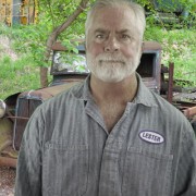 Man with gray hair and beard, wearing coveralls and standing in front of a rusted antique vehicle